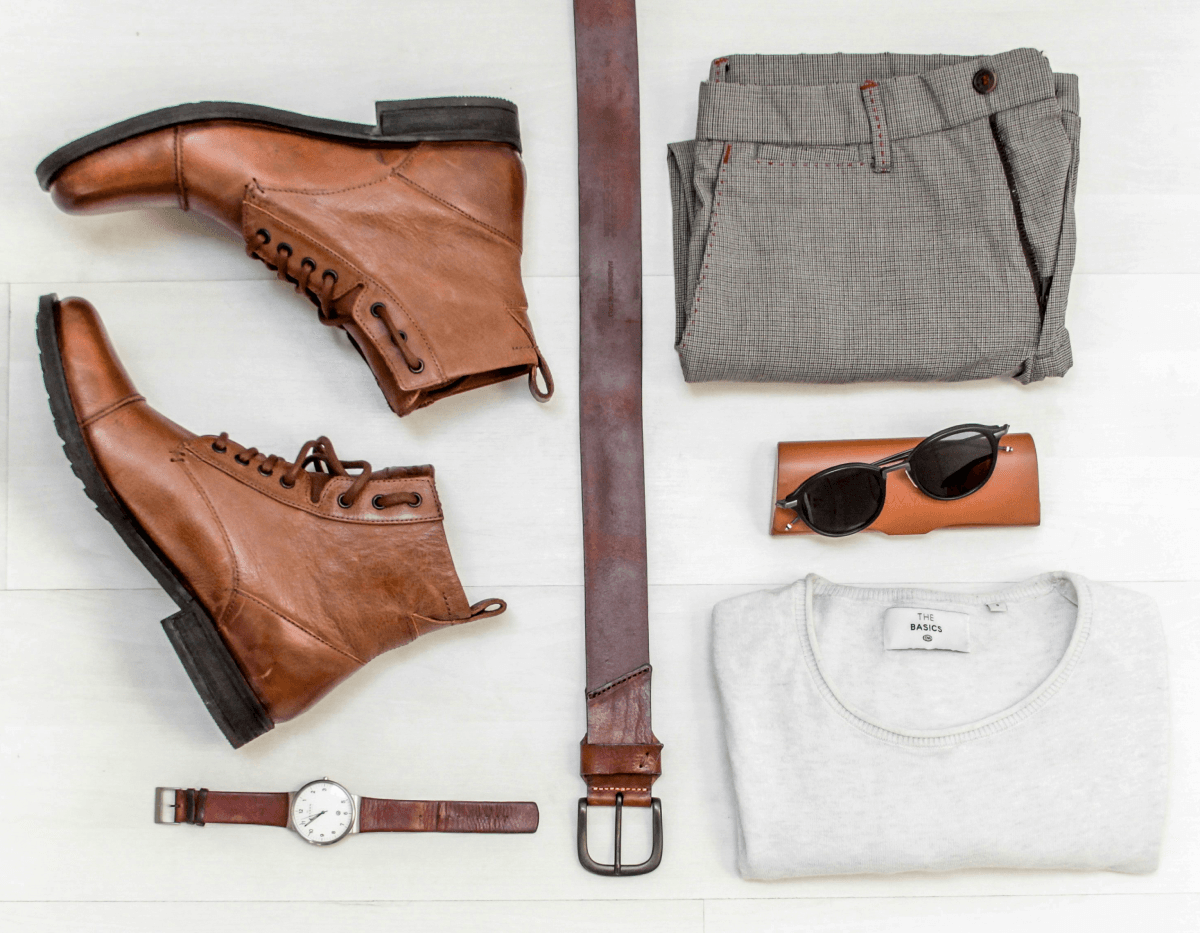 Flat lay of casual men's outfit with boots, belt, watch, sunglasses, sweater, and trousers