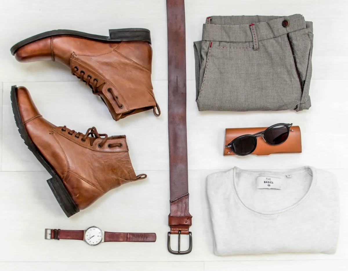 Flat lay of casual men's outfit with boots, belt, watch, sunglasses, sweater, and trousers