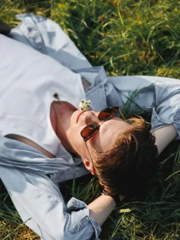 Man lying on grass in sunglasses, white tee, and light button-up shirt – relaxed. Displaying smart casual for men in a spring look