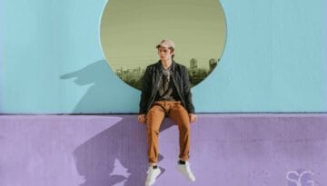 Young man in a casual spring outfit with brown pants, sneakers, and a cap, sitting against a pastel wall with circular cutout