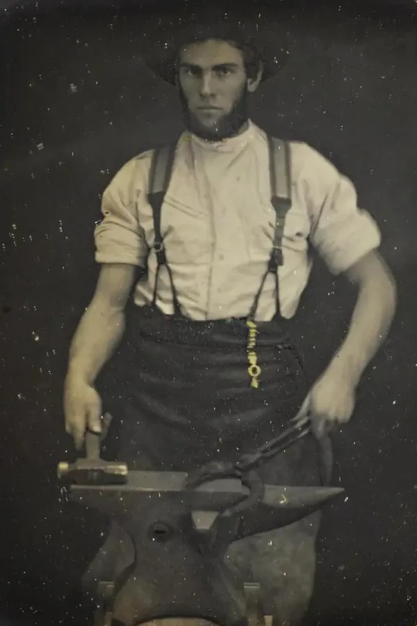 Vintage photo of a blacksmith wearing a white shirt, dark trousers, and leather suspenders while working at an anvil.