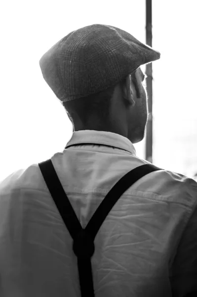 Man wearing Y-back leather suspenders and a flat cap, seen from behind in black and white photography.