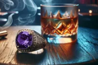 Vintage-style men’s ring with a large purple gemstone beside a glass of whiskey on a wooden table.