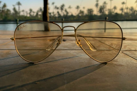 Gold aviator sunglasses resting on a wooden surface with palm trees and water in the background at sunset.
