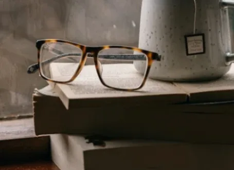 Tortoiseshell Wayfarer-style glasses resting on an open book next to a ceramic mug by a window.