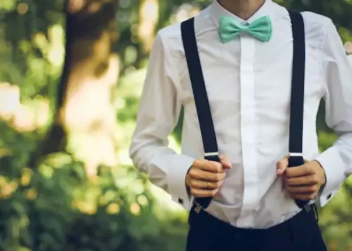 A man wearing leather suspenders and a mint green bow tie over a white shirt in an outdoor setting.