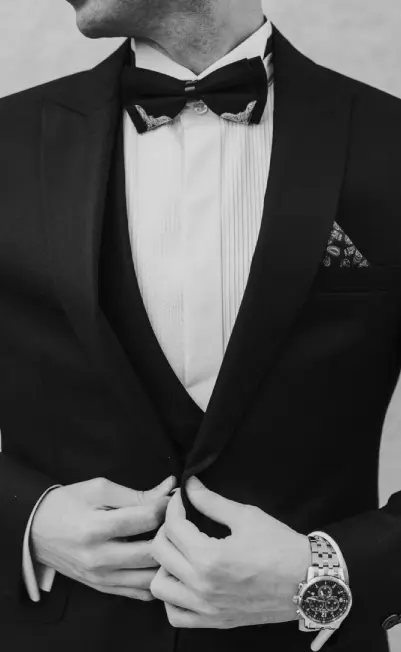 smart casual wear for men _ btc A man buttoning his tuxedo jacket with a watch and bow tie, showcasing smart casual wear for men.