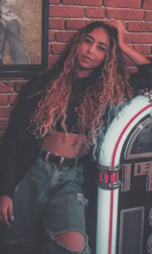 Woman in high-waisted ripped jeans and a crop top leans against a jukebox, styled in men's 90s clothing fashion.