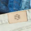 Close-up of light wash men's Spring denim jeans with an SG leather patch and stacked denim styles in the background.