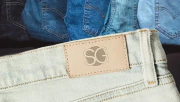 Close-up of light wash men's Spring denim jeans with an SG leather patch and stacked denim styles in the background.