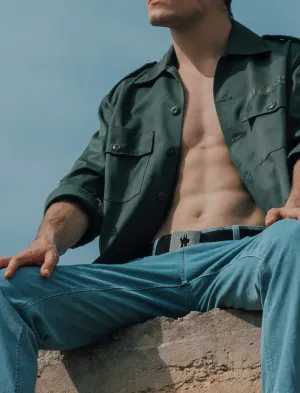 Man wearing an open dark green shirt with blue pants while sitting outdoors