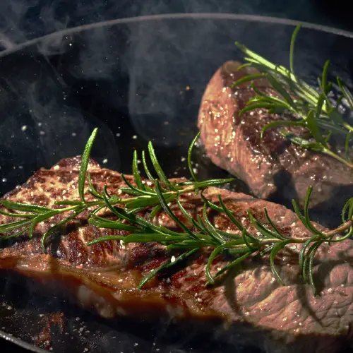 Cast iron pan searing steak with fresh rosemary, showing the kind of surface often recommended as the Best Pan for Steak.