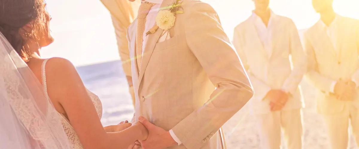 Man wearing a light beige summer suit during an outdoor beach wedding ceremony at sunset.