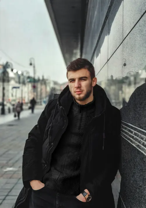 Man wearing an all black outfit with a black knit sweater, black pants, and a layered black jacket.
