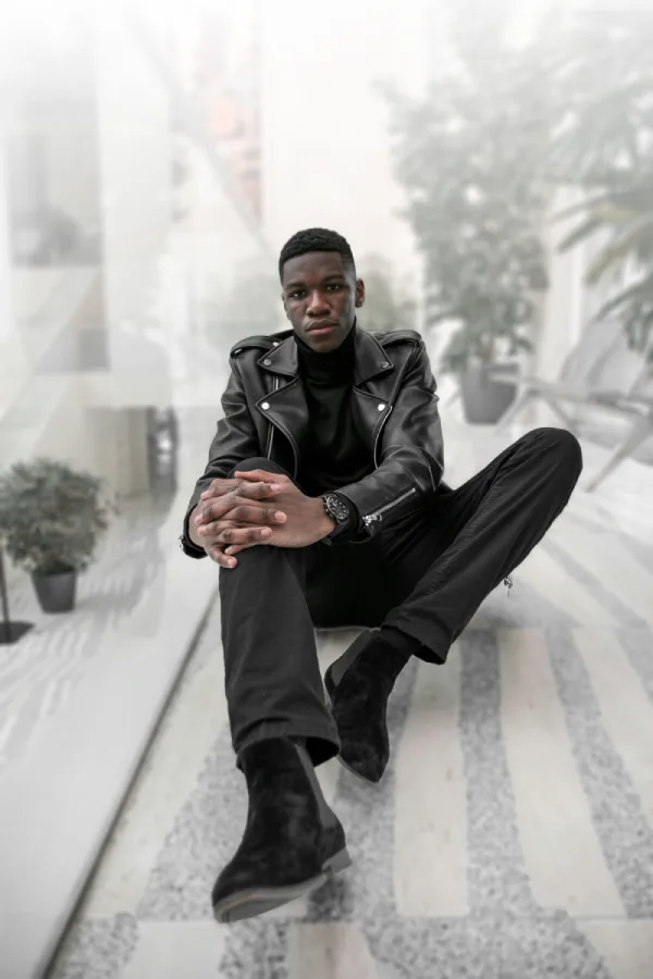 Man wearing an all black outfit with a black leather biker jacket, black turtleneck, black pants, and suede boots.