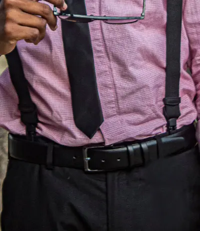 Close-up of a man wearing black suspenders and a black belt with dress trousers, showing why you do not wear a belt with suspenders.